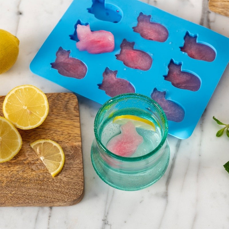 Blauwe ijsblokjesvorm op marmer met roze kattenvormige ijsblokjes, naast een glas water met een kattenblokje en citroen op een houten plank