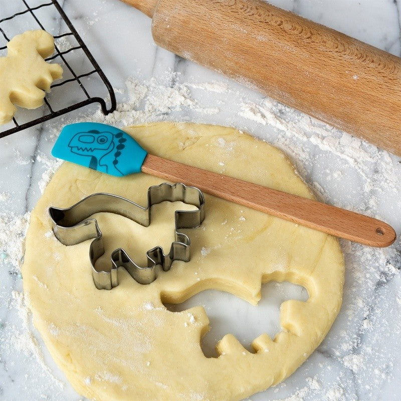 Dinovormige uitsteekvorm en turquoise spatel met houten handvat op uitgerold koekjesdeeg, naast een deegroller en een afkoelrek
