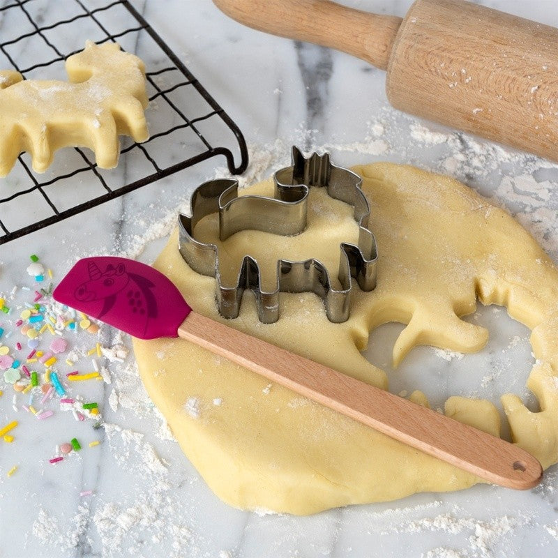 Roestvrijstalen eenhoornvorm in uitgerold koekjesdeeg op een marmeren blad, met de roze siliconen spatel, een houten deegroller, een koelrooster en sprinkles