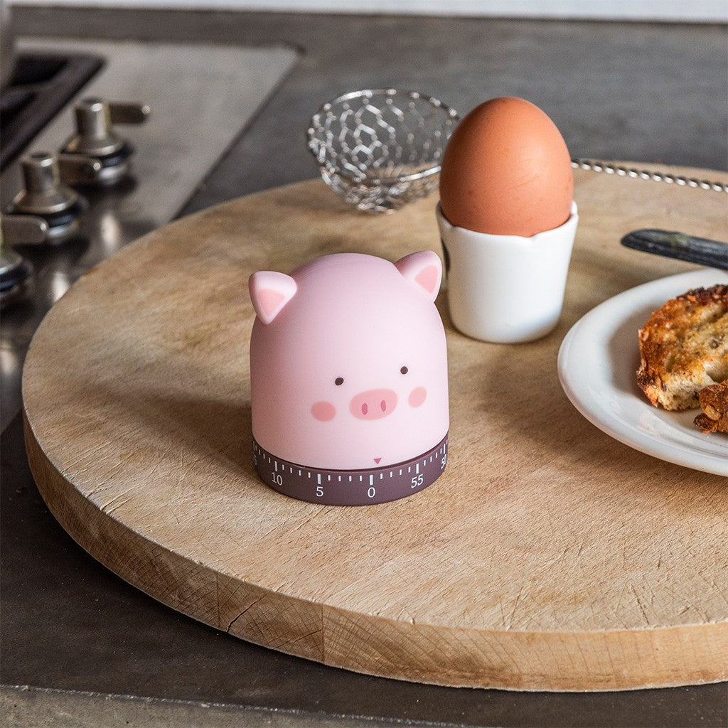 Roze varkenskookwekker op een ronde houten plank naast een eierdopje met een bruin ei en een bord met toast, bij het fornuis