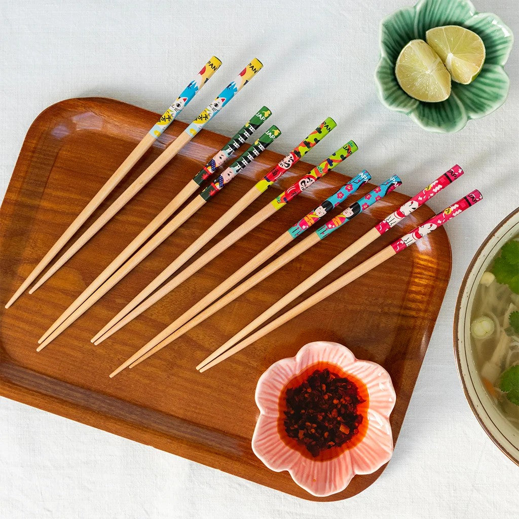 Vijf paar houten eetstokjes met kleurige katten en Japanse figuren op een houten dienblad, met een roze schaaltje chilliolie, een groen schaaltje met limoen en een kom soep
