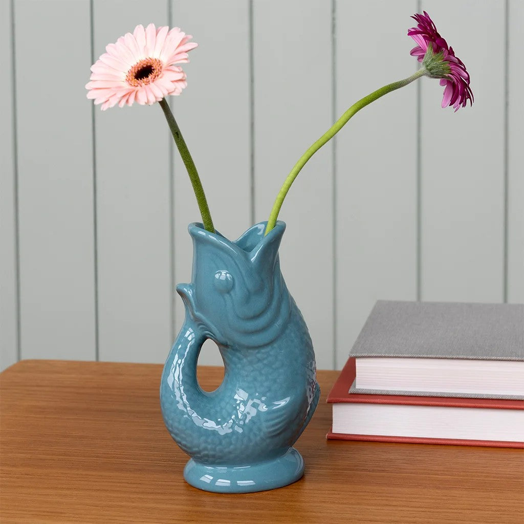Blauwe visvaas met twee gerbera's, een lichtroze en een paarsrode, op een houten tafel naast twee boeken voor een lichtgrijze plankenwand