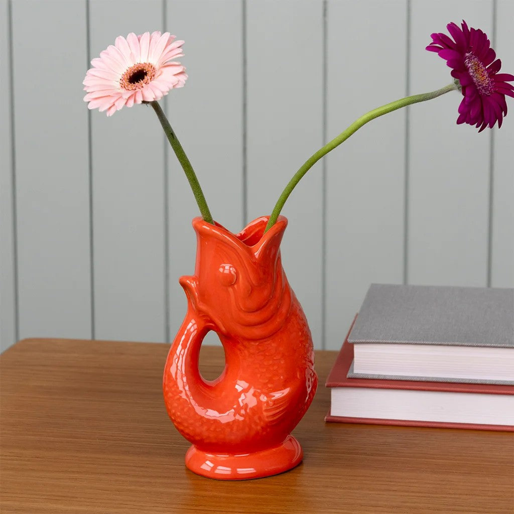 Oranje visvaas met een roze en een paarse gerbera op een houten tafel, naast een stapel boeken voor een grijsgroene plankenwand