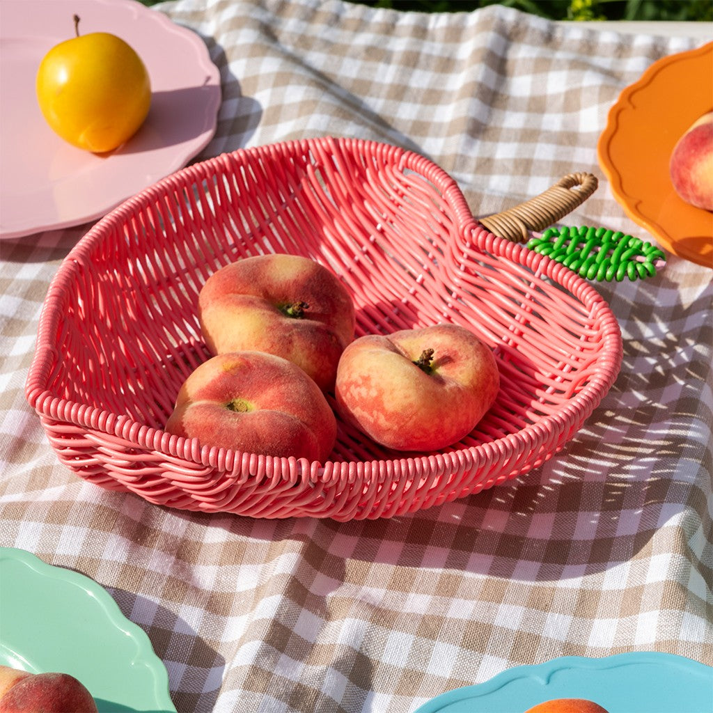 Roze gevlochten fruitmand met drie platte perziken op een geruit kleed tussen gekleurde borden
