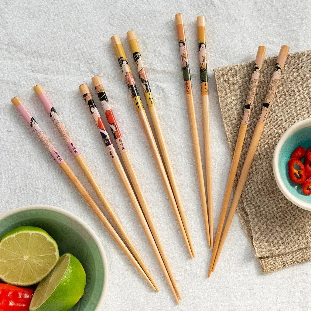 Vijf paar eetstokjes met geisha-portretten in roze, geel, oranje en beige op een wit linnen kleed, naast een groen schaaltje met limoen en een turquoise kommetje met gesneden rode peper