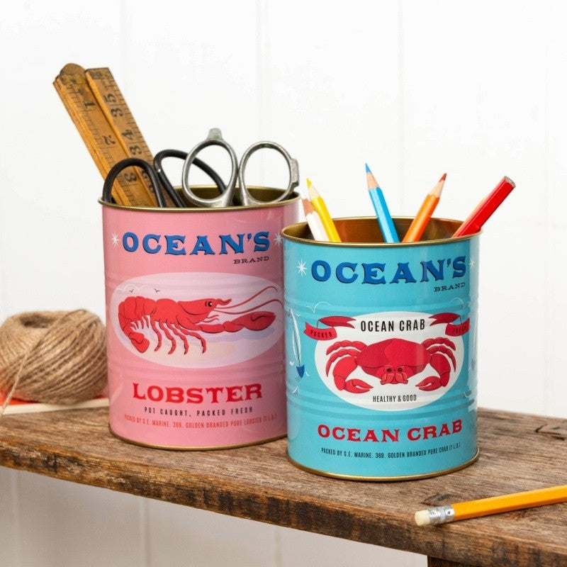 Roze kreeftblik met een duimstok en schaar en turquoise krabblik met kleurpotloden op een houten plank voor een witte wand
