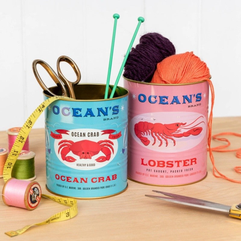 Turquoise krabblik met schaar en breinaalden en roze kreeftblik met bollen wol, naast een meetlint en garenklosjes op een houten tafel