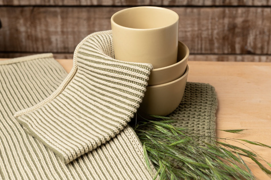 Solwang Design Keukendoek Rib - Olijfgroen sfeerfoto met de geribde keukendoek in olijfgroen, drie gestapelde kleine keramische kommetjes in zandbeige en een takje gedroogd gras op een houten ondergrond.
