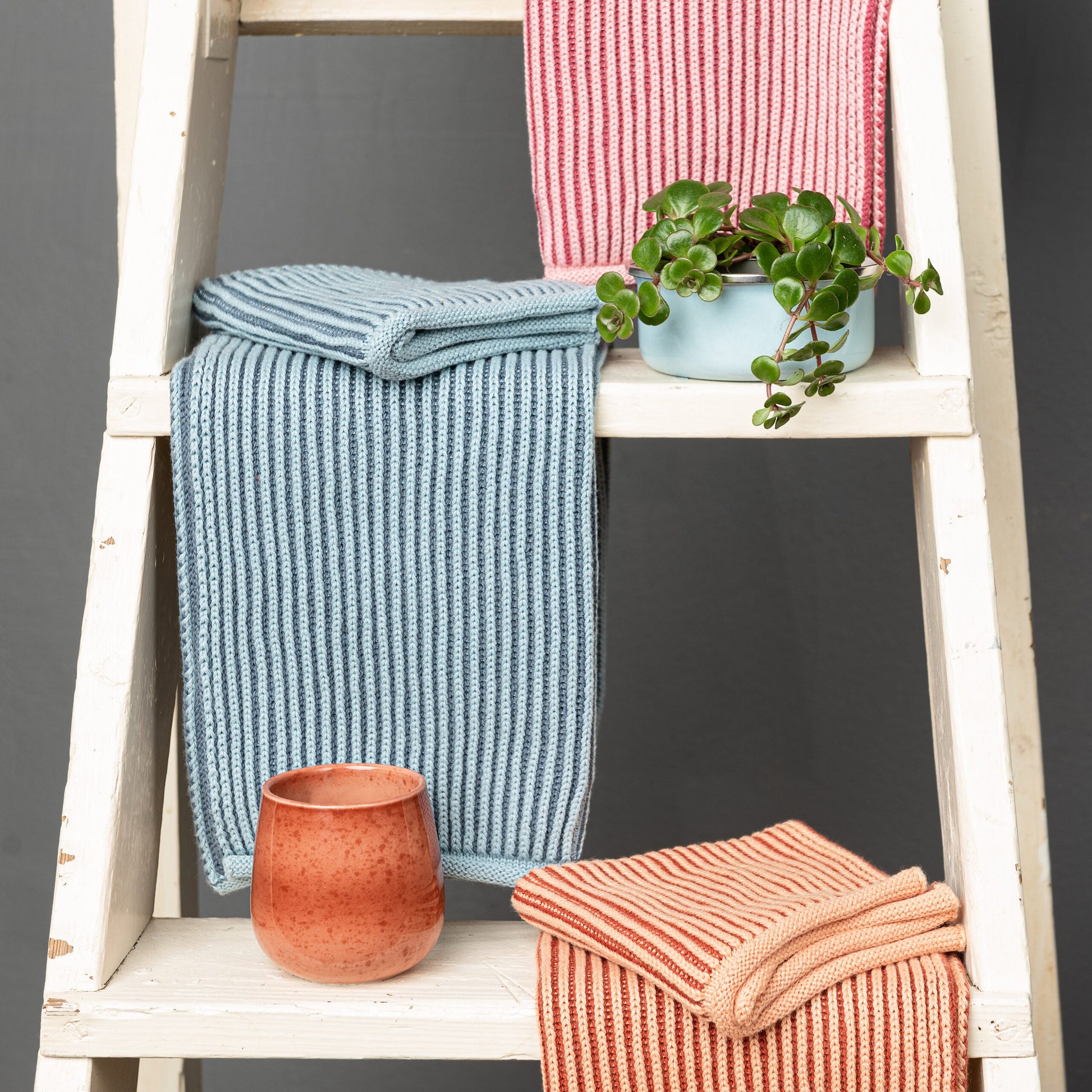 Solwang Design Keukendoek Rib - Blauwtinten sfeerfoto met de lichtblauwe geribde keukendoek gedrapeerd over een witte houten trapladder, op de achtergrond een roze en een oranje geribde keukendoek, een lichtblauw bakje met groene rankplant en een terracotta mok.