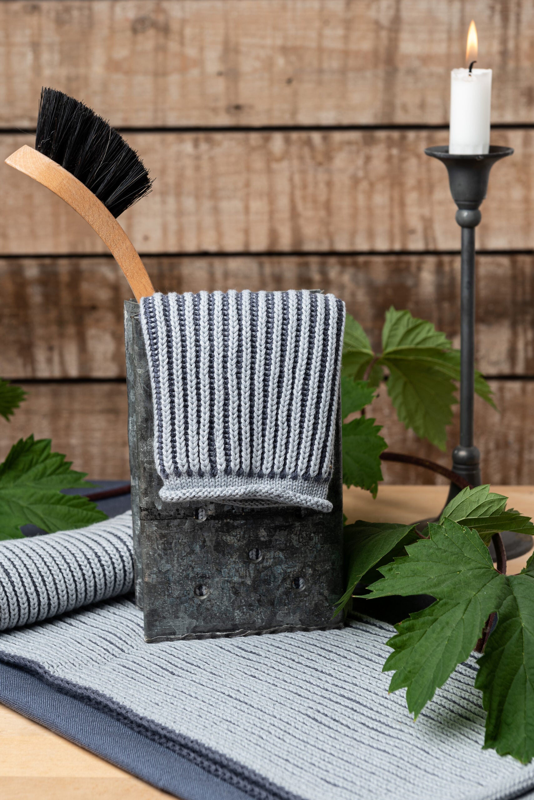 Solwang Design Keukendoek Rib - Stormy Grijs sfeerfoto met de grijze geribde keukendoek gedrapeerd over een verweerde zinken bak met daarin een houten afwasborstel, een brandende witte kaars op een zwarte kandelaar, groene wijnbladeren en een tweede gevouwen doek op tafel.