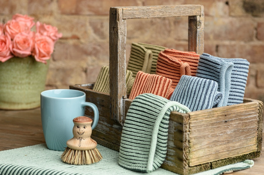 Solwang Design Keukendoek Rib - Petrol houten kist met daarin meerdere geribde keukendoeken in olijfgroen, terracotta, blauw en mintgroen, ernaast een lichtblauwe mok en een houten afwasborstel met geschilderd gezicht, op de achtergrond roze rozen in een vaas tegen een bakstenen muur.