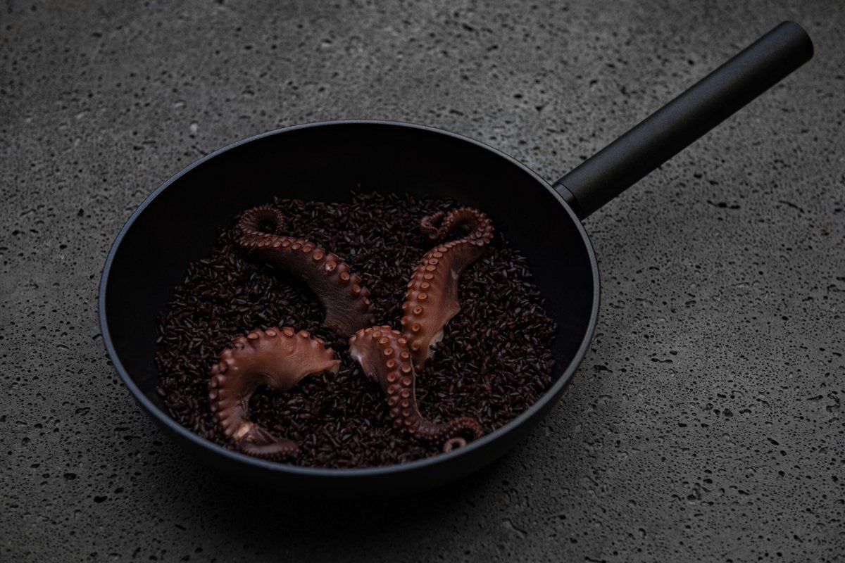 Zwarte wokpan gevuld met zwarte rijst en octopus op een donkere ondergrond