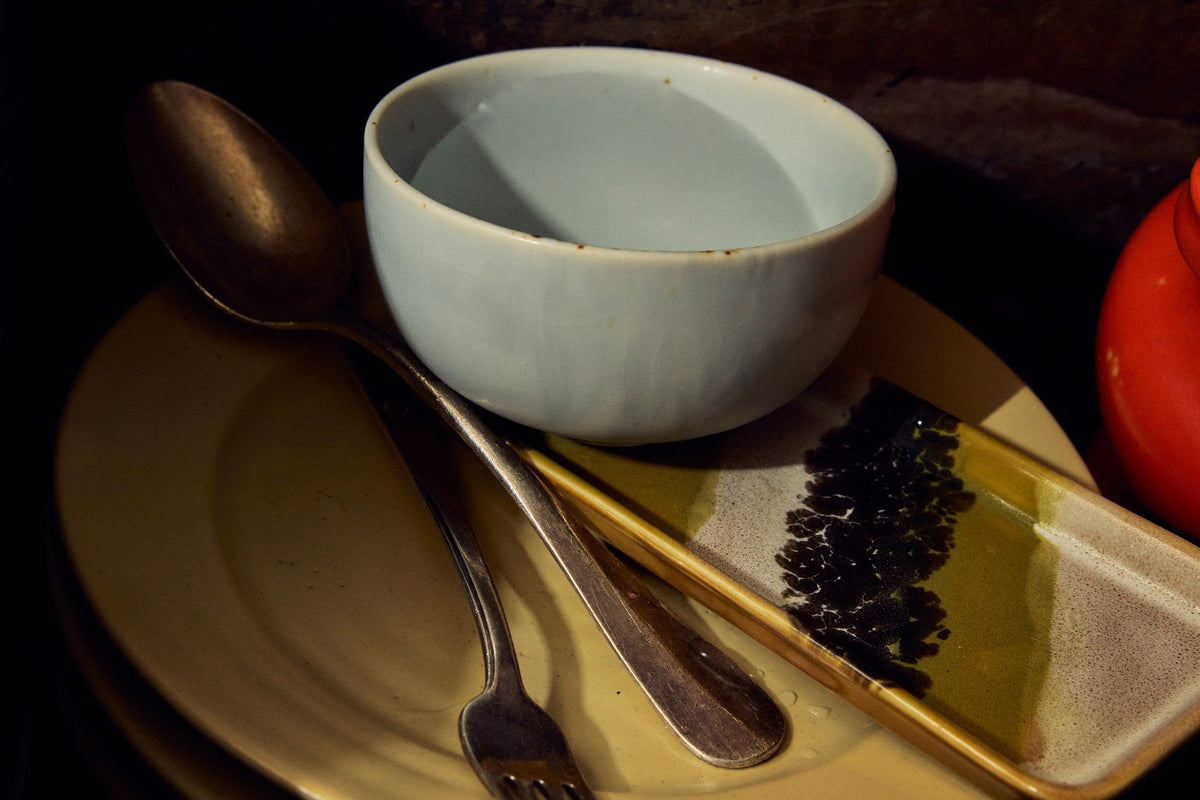 Chef Ceramics kom Soft Blue op een crèmekleurig bord naast een lepel, een vork en een rechthoekig schaaltje