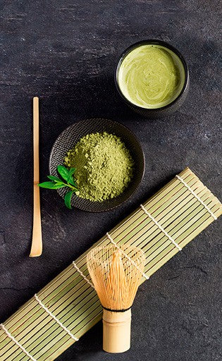 Ibili Matcha Set, sfeerbeeld met matchapoeder, een kom bereide matcha en de bamboe garde op een bamboe onderlegger.