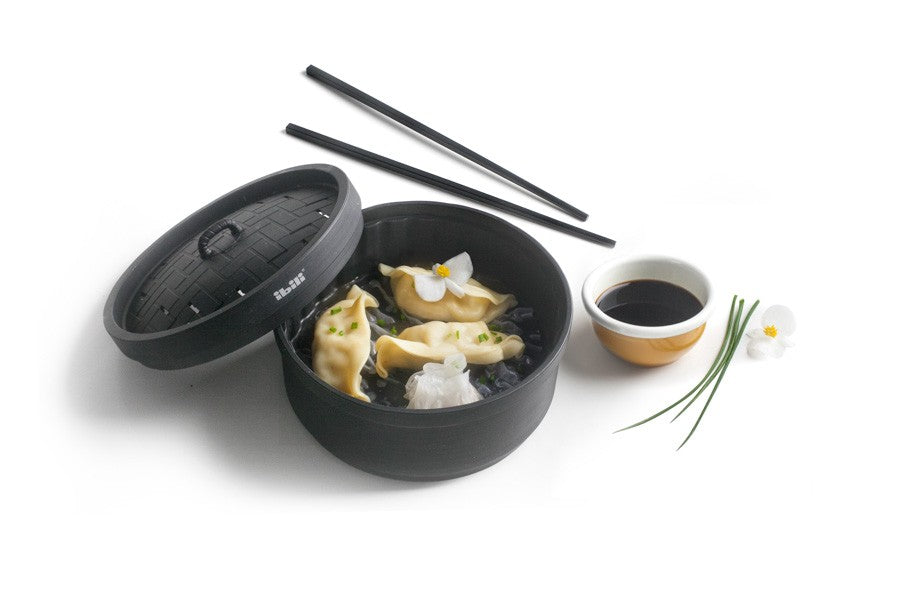 Ibili Stoommandje met Deksel zwart, gevuld met dumplings geserveerd met eetstokjes en een sausschaaltje.