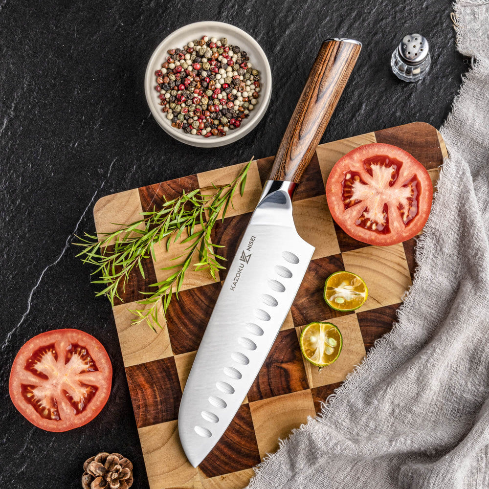 Kazoku Nisei santoku 18 cm op een snijplank met ruitpatroon naast tomaat, peperkorrels en rozemarijn, van bovenaf