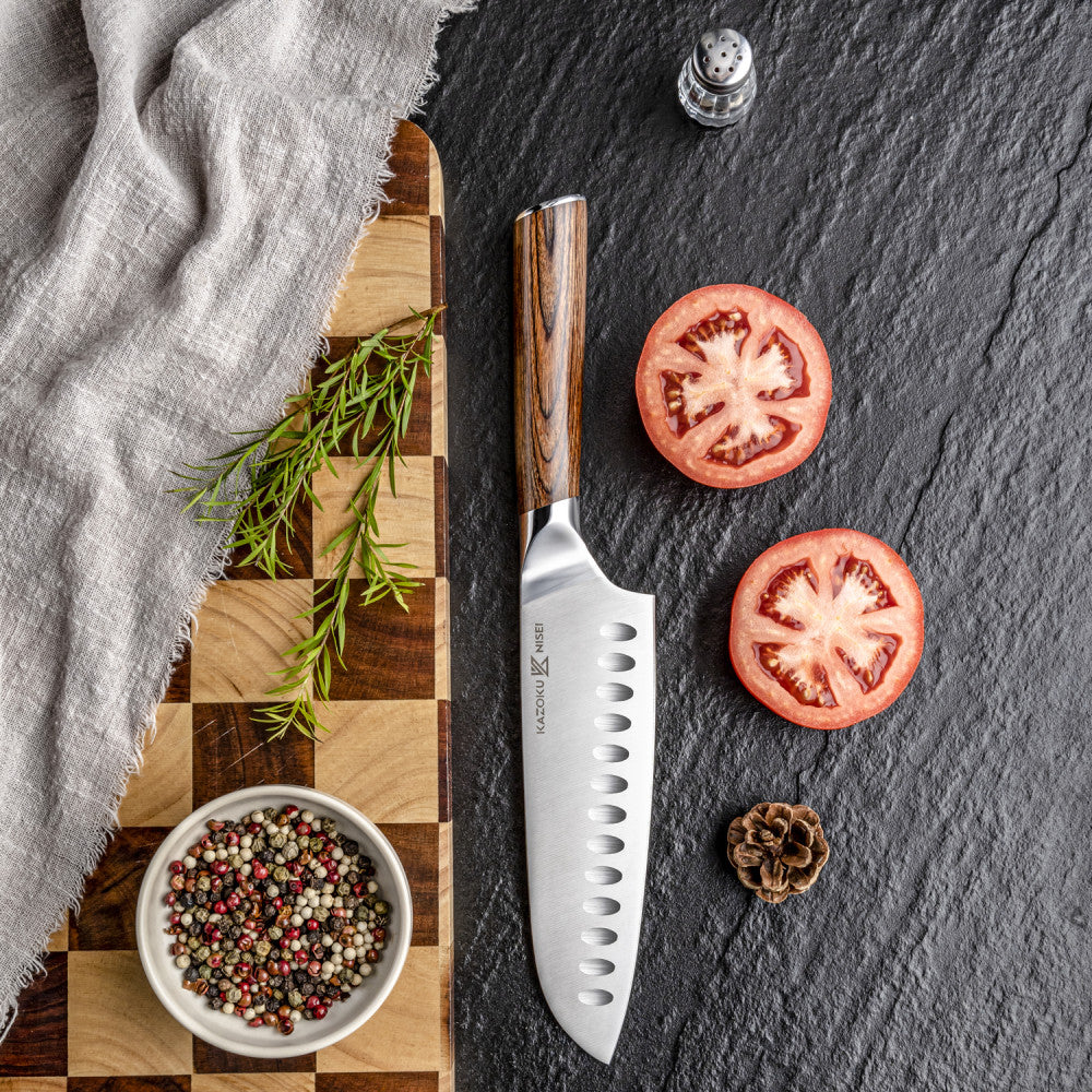 Kazoku Nisei santoku 18 cm rechtop op een snijplank met ruitpatroon naast tomaten en peperkorrels