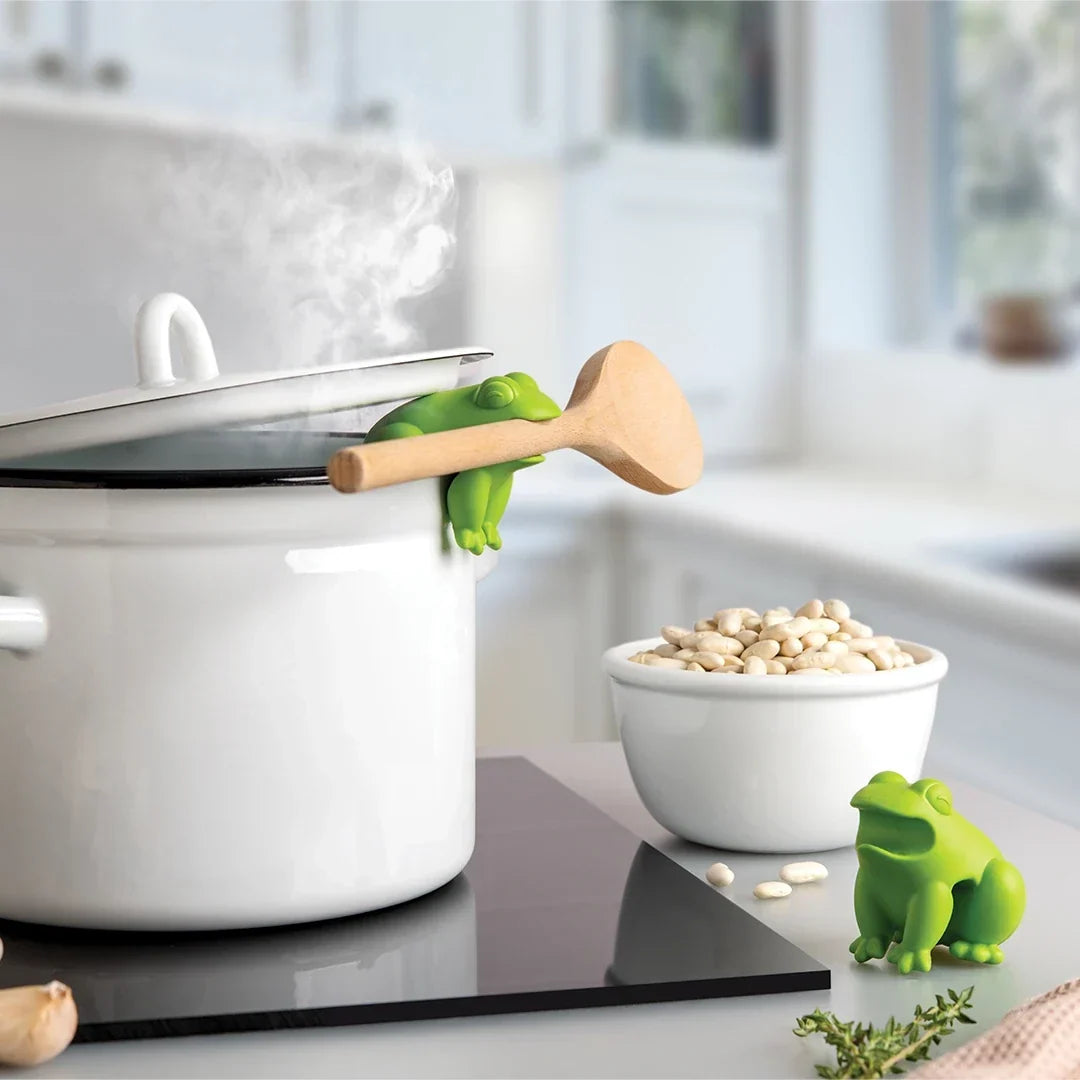 Groene kikker houdt een houten lepel vast op de rand van een pan