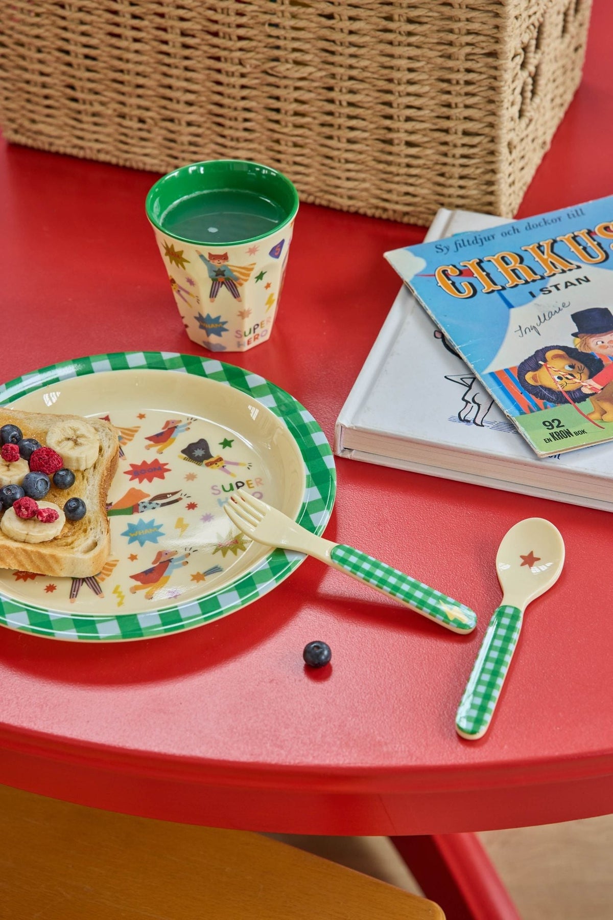 Kinderbeker met superheldenprint naast een kinderbord met boterham op een rode tafel