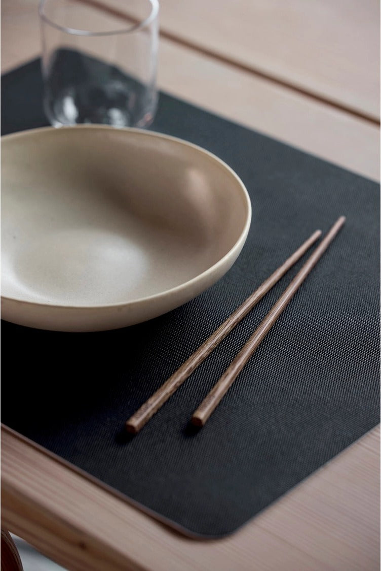 Shoku placemat Solid Black onder een beige kom, een glas en houten eetstokjes op een houten tafel