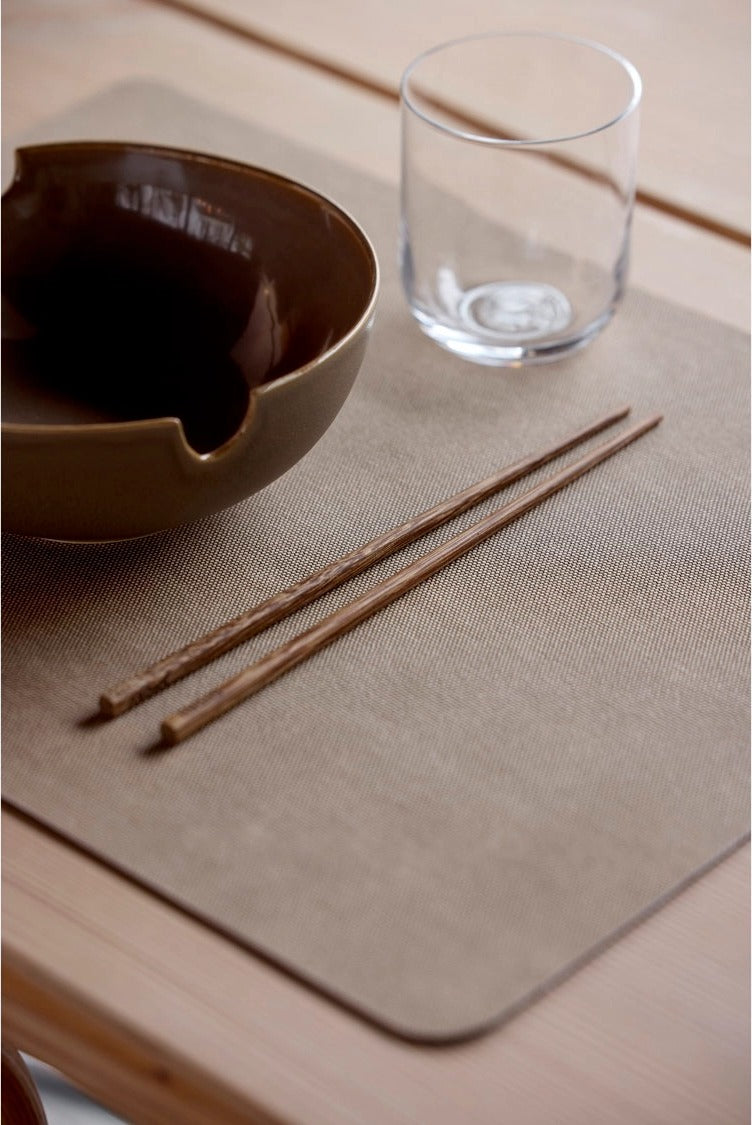 Shoku placemat Soft Beige met een donkerbruine kom, een glas en houten eetstokjes op een houten tafel